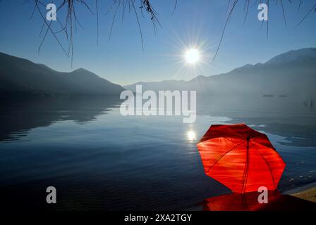 Parapluie rouge penché sur la plage de sable sur le lac alpin majeur avec montagne et Sunbeam et les îles Brissago dans une journée ensoleillée à Ascona, Tessin Banque D'Images