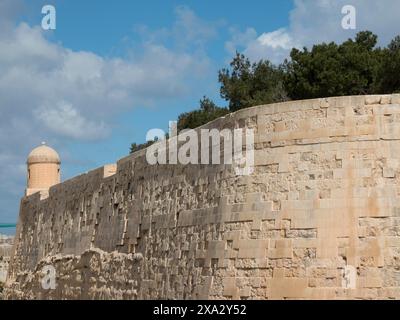 Mur massif de pierre d'une forteresse avec une tour et des arbres en arrière-plan sous un ciel bleu, Valette, Malte Banque D'Images