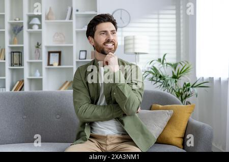 Homme barbu assis sur un canapé gris dans un salon élégant, perdu dans la pensée et le sourire. Intérieur de maison lumineux et confortable avec des plantes et un décor. Banque D'Images
