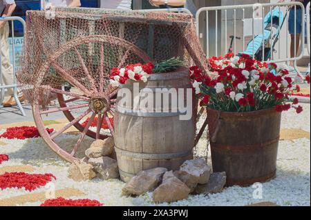 Scène rustique avec une roue de chariot en bois, un tonneau et un seau rempli de fleurs rouges et blanches sur un sol de pierres et de pétales. Banque D'Images