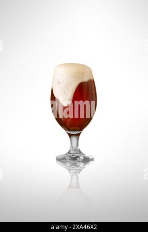 Photo de gobelet en verre débordant de bière artisanale sombre, surmonté d'une mousse épaisse et crémeuse sur fond blanc de studio. Banque D'Images