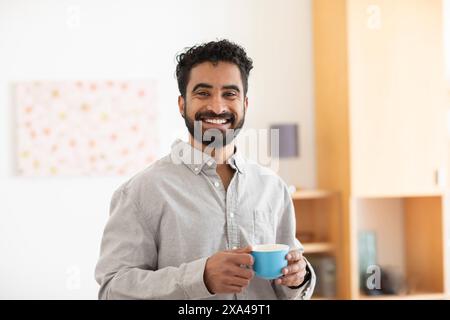 Un homme souriant avec une barbe tenant une tasse bleue se tient dans une pièce lumineuse avec des étagères Banque D'Images