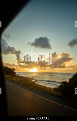 Une vue depuis une fenêtre de voiture capturant un magnifique coucher de soleil sur l'océan avec des nuages dispersés dans le ciel et un littoral sur la gauche. Banque D'Images