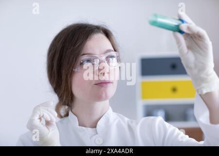Un scientifique concentré dans une blouse de laboratoire examine le contenu d'un tube à essai, portant des lunettes de sécurité et des gants. Banque D'Images