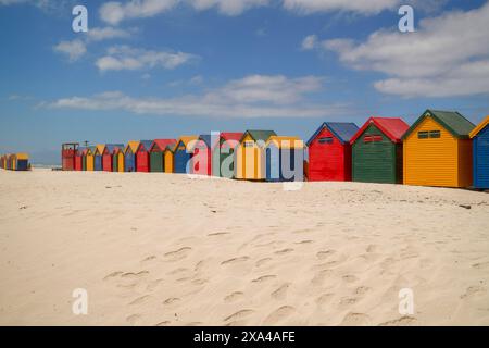 Une rangée de cabanes de plage colorées se dresse sur une plage de sable sous un ciel bleu clair. Banque D'Images
