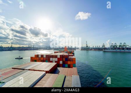 Un cargo est chargé de conteneurs colorés sous un ciel partiellement nuageux, naviguant près d'un port très fréquenté avec de nombreuses grues et des infrastructures maritimes. Banque D'Images