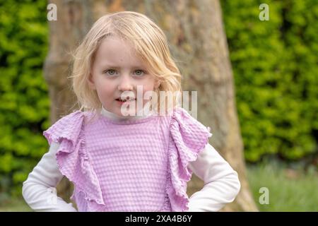 Une jeune fille aux cheveux blonds portant un haut tricoté violet se tient dehors avec un fond de verdure, souriant à la caméra. Banque D'Images