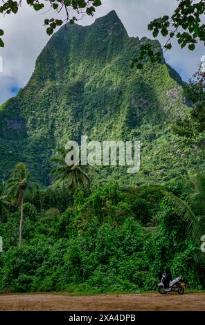 Une moto est garée sur un chemin de terre avec une végétation tropicale luxuriante et une montagne verdoyante en arrière-plan sous un ciel nuageux. Banque D'Images