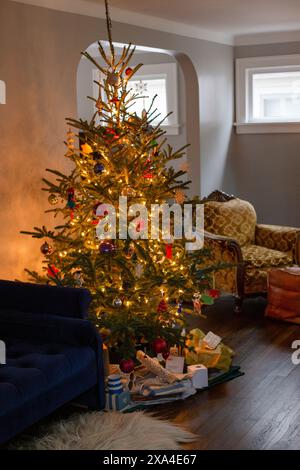 Un salon chaleureusement éclairé décoré pour Noël avec un sapin de Noël magnifiquement orné entouré de cadeaux emballés, et des meubles confortables en arrière-plan. Banque D'Images
