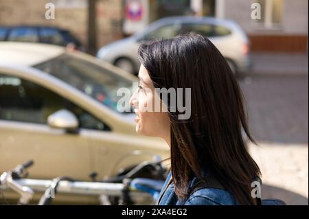 Une femme aux longs cheveux foncés est photographiée de profil, regardant vers l'avant par une journée ensoleillée, avec des voitures et des vélos en arrière-plan. Banque D'Images