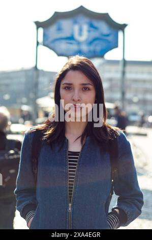 Une jeune femme se tient devant un fond de ville flou avec la lumière du soleil filtrant à travers, avec un panneau partiellement visible au-dessus de sa tête. Banque D'Images