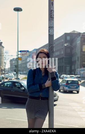 Une jeune femme se tient appuyée contre un bâton de rue dans un cadre urbain ensoleillé, portant une veste en Jean et une jupe rayée. Banque D'Images