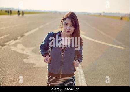 Une jeune femme se tient sur une route vide avec un arrière-plan doux, tenant sa veste ouverte avec confiance et regardant directement la caméra. Banque D'Images