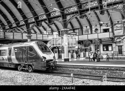 Un train repose sur un quai historique de la gare du XIXe siècle. Une passerelle est en arrière-plan et une femme attend sur un banc. Banque D'Images