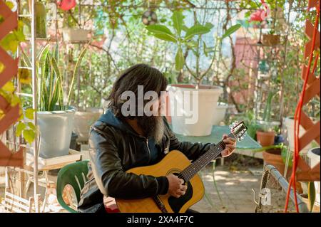 Un homme barbu aux cheveux longs joue de la guitare acoustique dans un jardin confortable entouré de plantes en pot et d'objets décoratifs. Banque D'Images