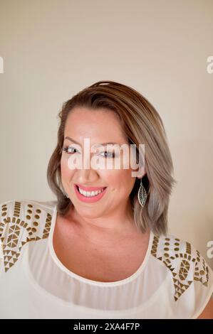 Une femme souriante avec des cheveux gris à la longueur de l'épaule, portant un haut blanc avec un motif d'épaule et des boucles d'oreilles en plumes, pose sur un fond beige clair. Banque D'Images