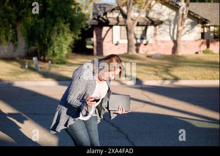 Une femme marche sur un trottoir alors qu'elle est engagée avec son smartphone et porte un ordinateur portable, avec en toile de fond des maisons résidentielles et des arbres. Banque D'Images