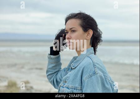 Une femme dans une veste en denim se tient à l'extérieur, avec un plan d'eau serein en arrière-plan, regardant au loin. Banque D'Images