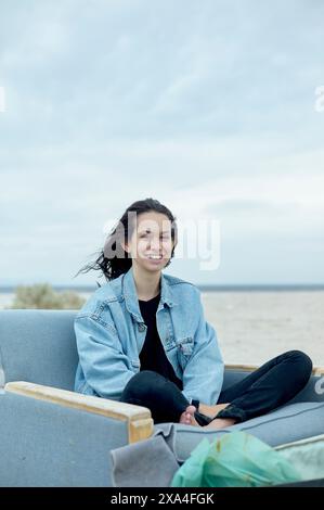 Une femme souriante s'assoit confortablement sur un canapé d'extérieur gris, portant une veste en denim et un pantalon noir, avec une plage de sable et un ciel nuageux en arrière-plan. Banque D'Images