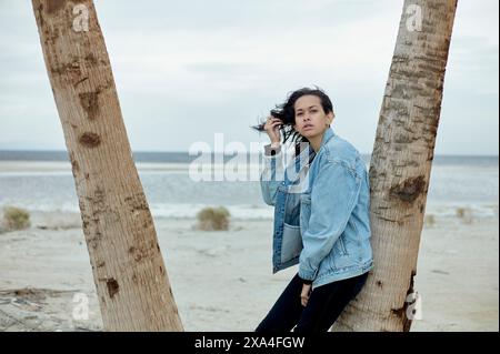 Une personne se tient entre deux palmiers sur une plage de sable, portant une veste en denim et un pantalon noir, regardant au loin avec une expression réfléchie. Banque D'Images