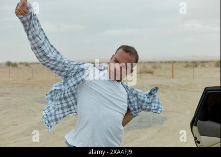 Un homme est à moitié à l'intérieur d'une voiture garée dans une zone désertique, enfilant énergiquement ou ajustant une chemise à carreaux bleus et blancs. Banque D'Images