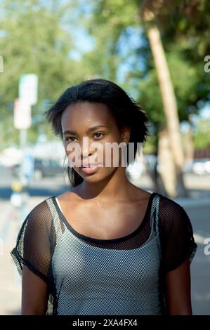 Une femme se tient debout dehors par une journée ensoleillée, avec des arbres et des panneaux de rue en arrière-plan, regardant la caméra avec une expression détendue. Banque D'Images