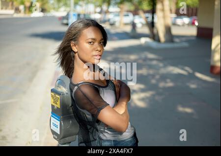 Une femme confiante se tient dans une rue ensoleillée avec les bras croisés, portant un top en maille noire et un sac à dos, regardant par-dessus son épaule la caméra. Banque D'Images