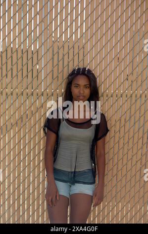 Une jeune femme se tient devant une clôture métallique, portant un haut noir transparent sur un vêtement plus léger et un short en Jean, avec ses cheveux partiellement vers le haut. Banque D'Images