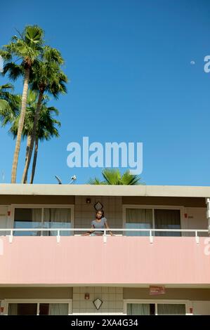 Une personne s'appuie sur la balustrade d'un balcon rose sous un ciel bleu clair, flanqué de palmiers et d'une mouette volante. Banque D'Images