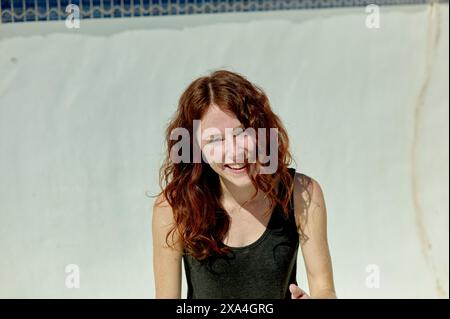 Une jeune femme joyeuse aux longs cheveux roux se tient près d'une piscine, son sourire rayonnant suggérant un moment de joie ou de rire en plein soleil. Banque D'Images