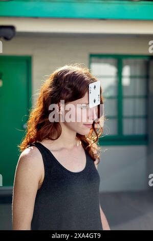 Une femme aux cheveux roux a une carte as de pique équilibrée sur son front ; elle se tient dehors avec un bâtiment vert en arrière-plan. Banque D'Images