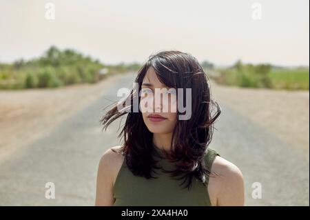 Une femme se tient sur une route déserte avec ses cheveux soufflant dans le vent, une expression douce et contemplative sur son visage. Banque D'Images