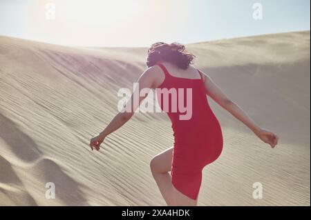 Une femme vêtue d'une robe rouge marche vers le haut d'une dune de sable sous la lumière du soleil. Banque D'Images