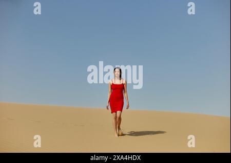 Une femme en robe rouge marche seule sur une dune de sable du désert sous un ciel bleu clair. Banque D'Images