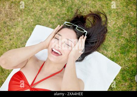 Une femme en maillot de bain rouge est allongée sur une serviette blanche sur l'herbe, riant avec ses mains sur sa tête et ses lunettes de soleil reposant au-dessus de son front. Banque D'Images