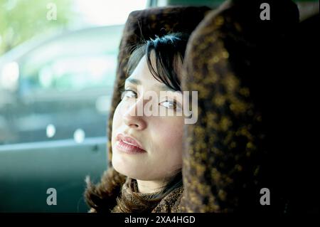 Une femme regarde soigneusement par la fenêtre d'une voiture, ses yeux scintillant contre une lumière naturelle douce, soulignant son expression contemplative. Banque D'Images