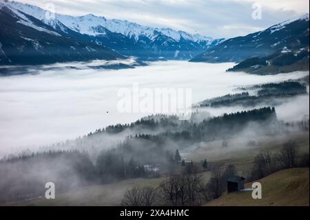 Un paysage montagneux brumeux avec des couches de brouillard installées dans les vallées, une petite cabane au premier plan et des sommets enneigés en arrière-plan. Banque D'Images