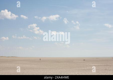 L'image montre une vaste plage de sable avec un ciel bleu calme parsemé de nuages blancs. Les gens apparaissent comme de minuscules figures au loin, soulignant la nature expansive du cadre de plage. Banque D'Images