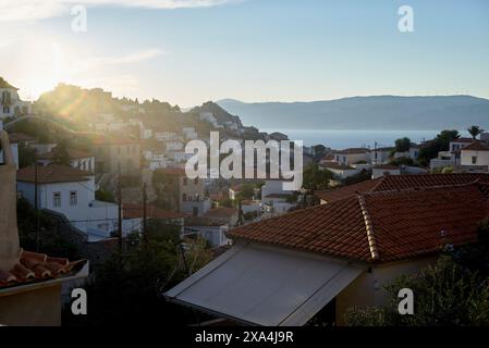 Une vue panoramique d'une ville côtière au lever du soleil avec des toits en terre cuite et le soleil jetant un coup d'œil sur la colline, jetant une lumière douce sur les bâtiments et la mer calme en arrière-plan. Banque D'Images