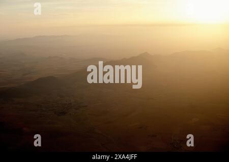 Vue aérienne d'un paysage montagneux baigné dans la lueur chaude du coucher ou du soleil levant, avec une brume diffusant la lumière du soleil. Banque D'Images