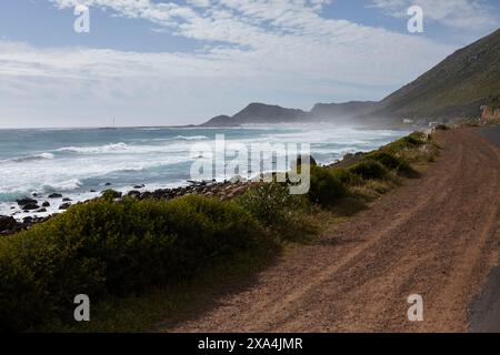 Les vagues de l'océan s'écrasent contre un rivage rocheux parallèle à un chemin de terre, avec un fond montagneux partiellement enveloppé par la brume. Banque D'Images