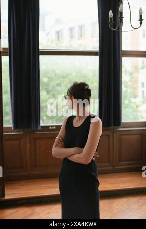 Une femme se tient près d'une fenêtre avec les bras croisés, regardant à l'extérieur d'une pièce avec d'élégants lambris en bois et des rideaux sombres. Banque D'Images