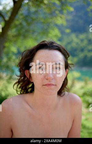 Portrait d'une jeune femme avec des taches de rousseur, des cheveux bruns et une expression neutre, debout à l'extérieur avec un fond naturel flou. Banque D'Images