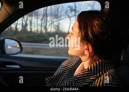 Une femme est assise à l'intérieur d'une voiture regardant par la fenêtre du passager pendant un coucher de soleil, avec la lumière douce du soleil illuminant la moitié de son visage. Banque D'Images