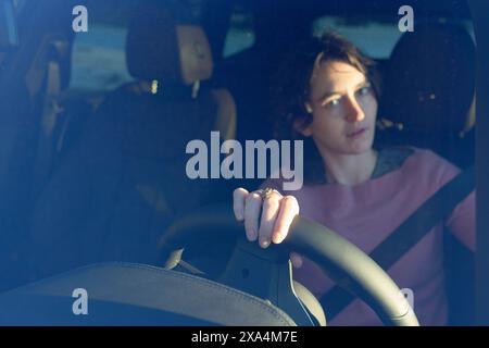 Une femme est assise derrière le volant dans un véhicule, regardant pensive quand elle regarde par la vitre du côté conducteur, avec le soleil projetant des ombres à travers l'intérieur. Banque D'Images