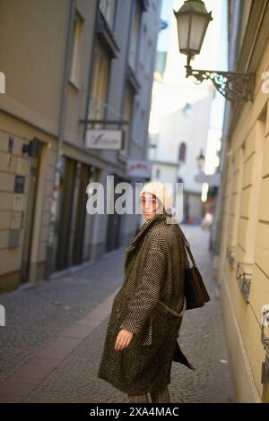 Une femme portant un manteau à pied de poule à la mode et un béret blanc regarde par-dessus son épaule tout en marchant dans une rue étroite à l'architecture européenne. Banque D'Images