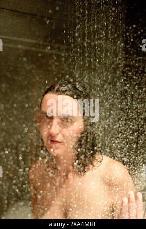 Une jeune femme se tient sous une douche avec des gouttelettes d'eau en cascade vers le bas, des cheveux mouillés plâtrés sur leur tête, et une expression neutre sur leur visage. Banque D'Images