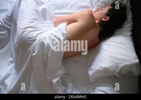 Une jeune femme est vue dormir paisiblement sur un lit recouvert de draps blancs, le dos partiellement exposé et la tête reposant sur un oreiller blanc. Banque D'Images