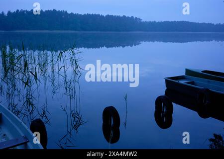 Une scène tôt le matin à un lac calme avec un reflet clair des arbres à la surface de l'eau et des bateaux au bord. Banque D'Images