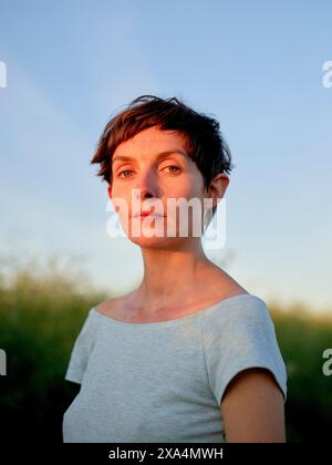 Une femme aux cheveux courts se tient dehors pendant l'heure dorée, son regard dirigé légèrement hors caméra. Banque D'Images